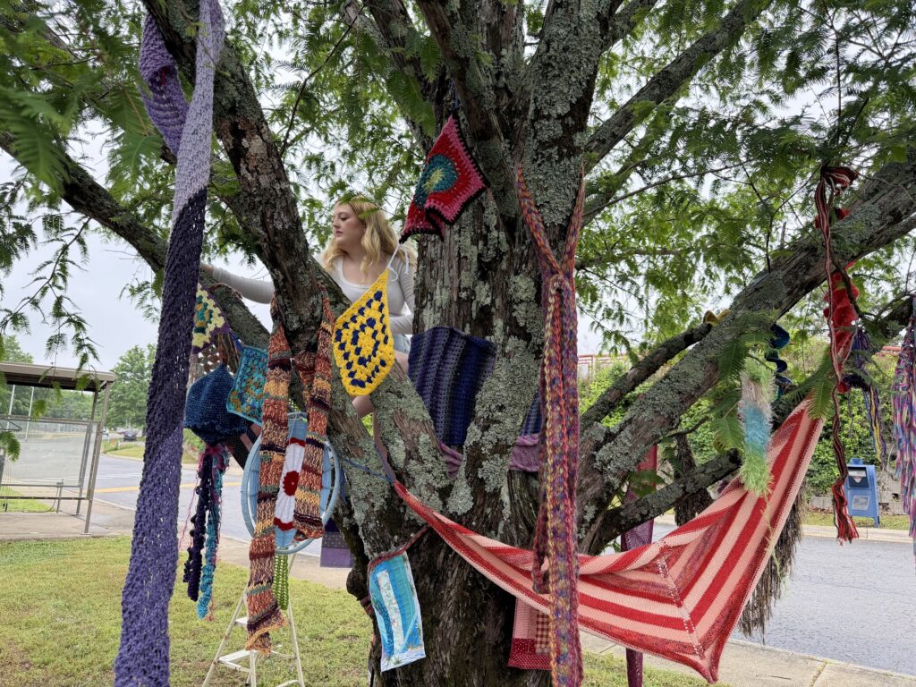 Person hangs hand-made crocheted and knitted adornments in a tree outside the library.